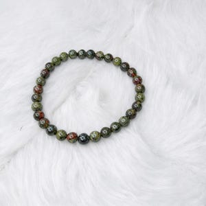 May include: A circular beaded bracelet featuring polished, spherical stones in mottled shades of forest green, olive, and deep reddish-brown. The beads are uniform in size and strung together to form a continuous loop, showcasing natural color variations.