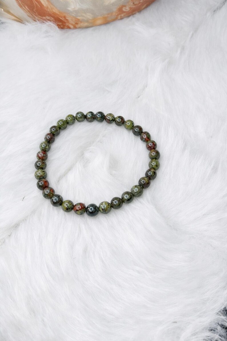 May include: A circular beaded bracelet featuring polished, spherical stones in mottled shades of forest green, olive, and deep reddish-brown. The beads are uniform in size and strung together to form a continuous loop, showcasing natural color variations.