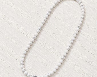 May include: A single strand necklace composed of small, round, white beads with grey veining patterns. The beads are uniform in size and arranged in a continuous circular loop, creating a simple, clean aesthetic with a single darker grey bead.