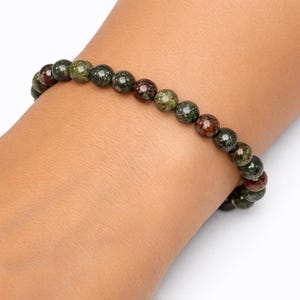 May include: A beaded bracelet featuring round, polished stones in mottled shades of forest green, olive, and deep reddish-brown. The beads are strung together in a single continuous loop, creating a uniform, circular design that rests on the wrist.