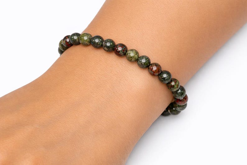 May include: A beaded bracelet featuring round, polished stones in mottled shades of forest green, olive, and deep reddish-brown. The beads are strung together in a single continuous loop, creating a uniform, circular design that rests on the wrist.