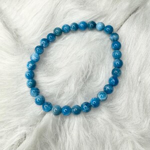 Puede incluir: Una pulsera de cuentas azules. La pulsera est&aacute; hecha de cuentas redondas de color azul claro ensartadas en un cord&oacute;n el&aacute;stico transparente. Las cuentas tienen un aspecto ligeramente veteado. La pulsera se exhibe sobre una superficie blanca y esponjosa.