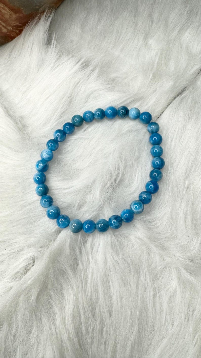 Puede incluir: Una pulsera de cuentas azules. La pulsera est&aacute; hecha de cuentas redondas de color azul claro ensartadas en un cord&oacute;n el&aacute;stico transparente. Las cuentas tienen un aspecto ligeramente veteado. La pulsera se exhibe sobre una superficie blanca y esponjosa.