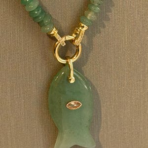 May include: A green beaded necklace with a gold clasp and a fish-shaped pendant. The pendant has a gold-colored accent in the center. The necklace is displayed on a neutral background.