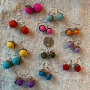 May include: A collection of handmade acorn earrings in various colors, including pink, orange, yellow, teal, purple, and red. Each earring features a felt acorn with a brown cap and silver-tone ear wires. The earrings are displayed on a neutral linen surface.