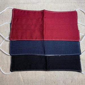 Puede incluir: Tres mascarillas faciales apiladas, en rojo, azul marino y negro. Cada mascarilla es rectangular, con costuras blancas y gomas elásticas blancas para las orejas. Las mascarillas son de tela.