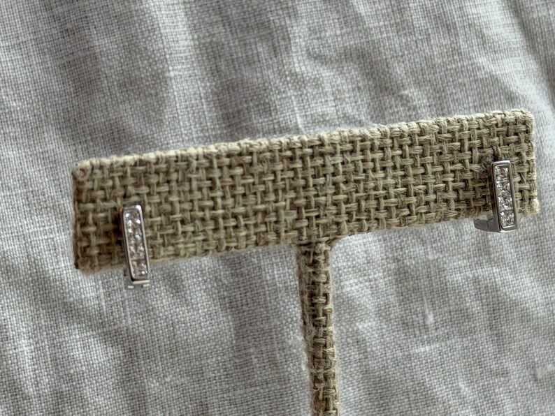May include: A pair of silver bar stud earrings, each adorned with a row of small, clear crystals. The earrings are displayed on a textured, tan jewelry display stand against a neutral background.