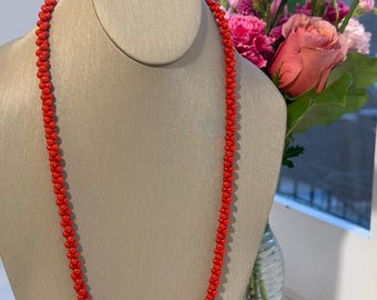 May include: A long, vibrant red beaded necklace displayed on a mannequin. The necklace is composed of small, round beads, creating a continuous strand. A bouquet of pink flowers sits in the background, adding a touch of color.