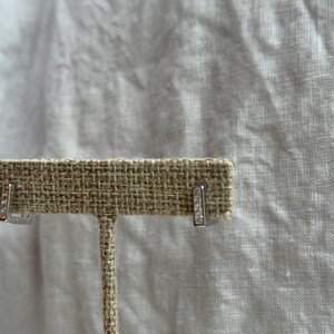 May include: A pair of silver-toned bar earrings with small, clear gemstones. The earrings are displayed on a tan, textured earring holder against a white background.