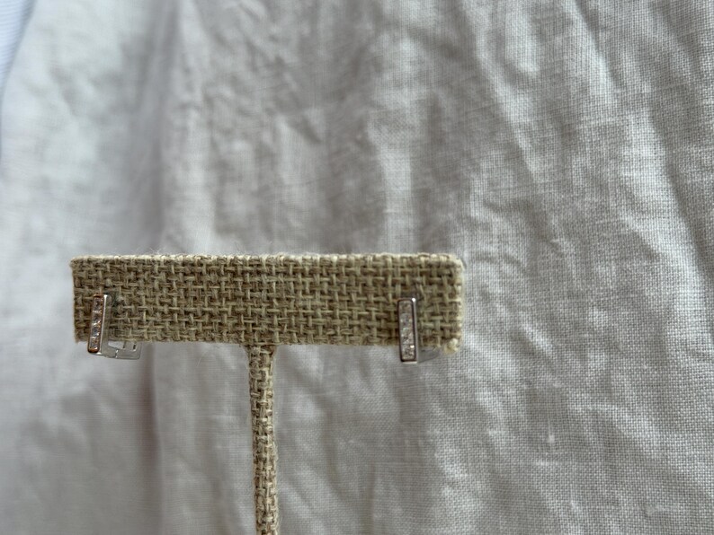 May include: A pair of silver-toned bar earrings with small, clear gemstones. The earrings are displayed on a tan, textured earring holder against a white background.