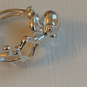 May include: A silver-colored octopus charm, with a detailed design, is attached to a ring. The octopus has a rounded head and eight tentacles. The charm is highly reflective, suggesting a polished metal finish. The background is a light, neutral color.
