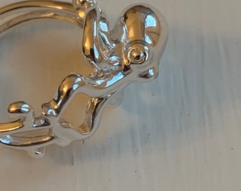 Peut inclure: Un charm argenté en forme de poulpe, avec un design détaillé, est fixé à un anneau. Le poulpe a une tête arrondie et huit tentacules. Le charme est très réfléchissant, suggérant une finition en métal poli. Le fond est de couleur claire et neutre.