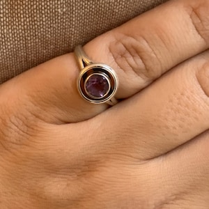 May include: A silver ring with a circular design, featuring a deep purple gemstone at the center. The ring's band splits into two graceful curves as it approaches the gemstone setting, creating a modern and elegant look. The ring is shown on a hand.