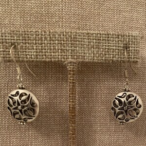 May include: A pair of silver-colored dangle earrings. Each earring features a round, openwork design with a floral pattern. The earrings hang from a simple hook and have a small bead detail at the top and bottom of the round design.