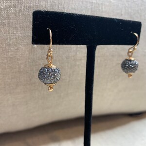 May include: Gold-toned drop earrings with a small, round, gray, crystal-covered bead. The earrings have a simple hook design and a small gold bead at the bottom of the crystal ball. The earrings are displayed on a black stand.