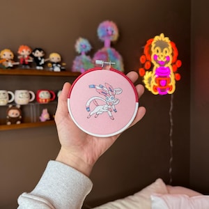 May include: A pink and red embroidery hoop with a white embroidered image of a Pokemon character, Sylveon, with pink and blue accents.