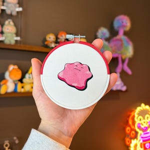 May include: A pink and white embroidered hoop with a pink blob-like character with a black outline. The hoop is held in a hand.