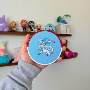 May include: A round embroidery hoop featuring a cartoon Sylveon design on light blue fabric. The hoop has a red and white border. The image is of a handmade craft, possibly for wall decor or a gift.