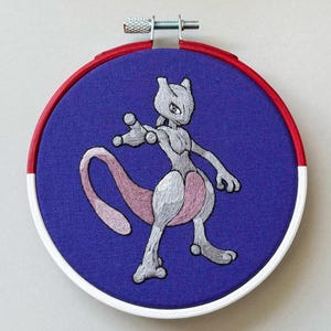 May include: Embroidered hoop art featuring a gray and pink Pokemon character, Mewtwo, on a purple background. The hoop is white with red and white trim.