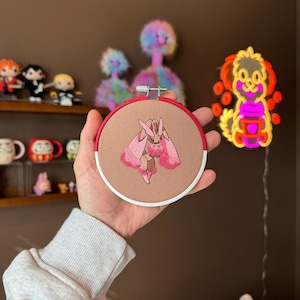 May include: A pink and red embroidered hoop art featuring a cartoon pink rabbit with long ears and a white fluffy tail.