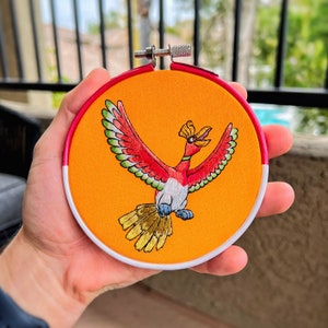 May include: Embroidered hoop art featuring a vibrant Ho-Oh design. The artwork showcases a colorful bird with red, green, and yellow details against an orange background. The hoop has a pink and white border, creating a decorative piece.