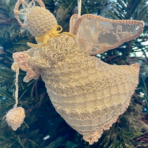 May include: A crocheted angel ornament with a white and pink color scheme. The angel has a halo, wings, and a beaded dress. The ornament is hanging from a tree branch.