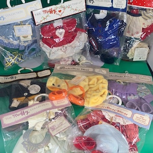May include: A collection of doll clothes packaged in clear plastic bags. The clothes are for a doll named "Muffy" and include a red dress with white trim, a blue shirt with a white collar, a purple dress with white trim, a yellow dress with white trim, a white dress with a white sash, and a red and white plaid dress with a white hat.