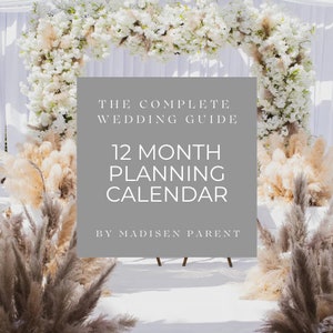 May include: A gray sign with white text that reads "The Complete Wedding Guide 12 Month Planning Calendar by Madisen Parent" against a backdrop of a white wedding arch decorated with white flowers and pampas grass.