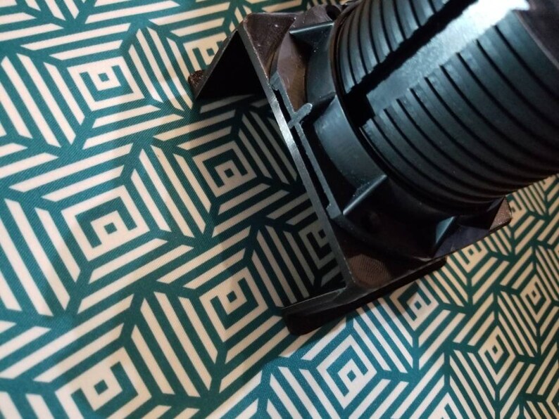 May include: Close-up of a black plastic electrical outlet cover with a ribbed design. The cover is on a white and teal patterned surface.