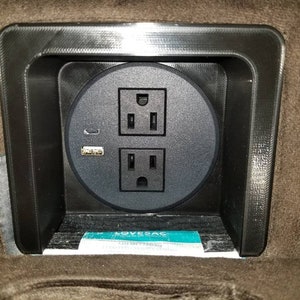 May include: A black electrical socket with two standard outlets and a USB port, mounted in a brown fabric surface.