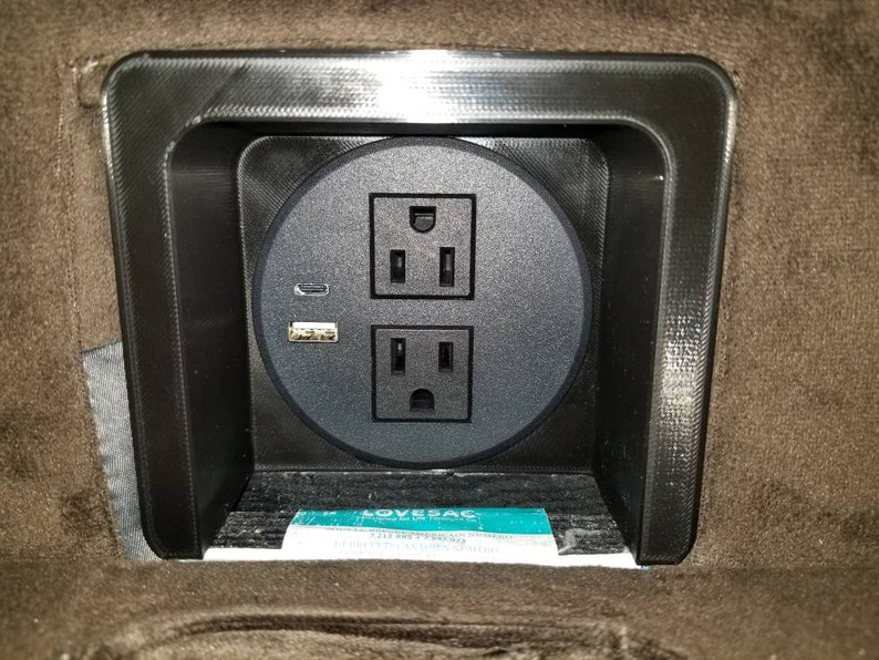 May include: A black electrical socket with two standard outlets and a USB port, mounted in a brown fabric surface.