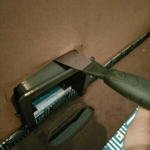May include: A black plastic storage compartment with a blue and white label is being opened with a black plastic scraper tool.