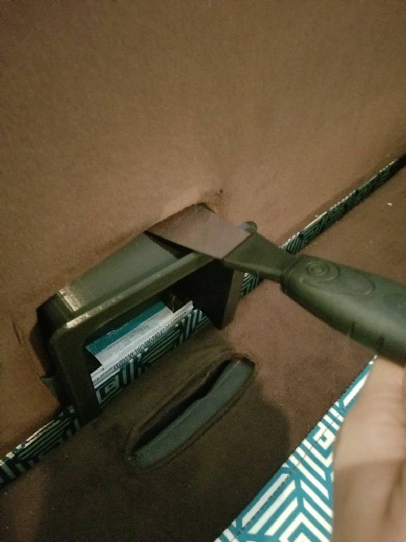 May include: A black plastic storage compartment with a blue and white label is being opened with a black plastic scraper tool.