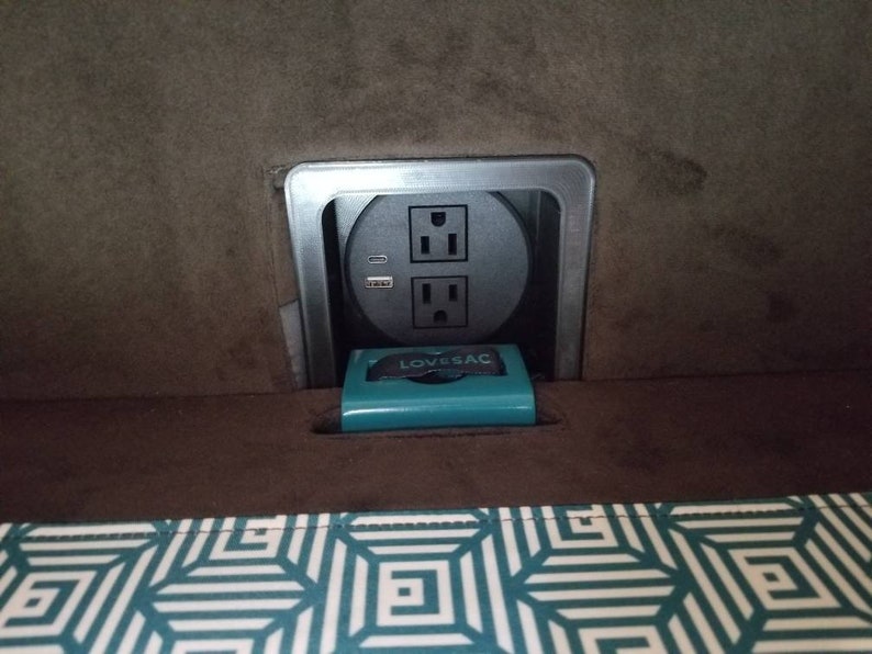 May include: A close-up of a power outlet with two standard sockets and a USB port. The outlet is mounted in a brown wall and is surrounded by a silver trim. A small teal box with the word "Lovesac" printed on it is sitting in front of the outlet.