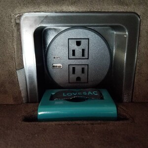 May include: A close-up of a grey electrical outlet with two standard sockets and a USB port. The outlet is mounted in a grey metal frame and is surrounded by a brown fabric surface. The text "LOVE SAC" is visible on a teal-coloured object in the foreground.