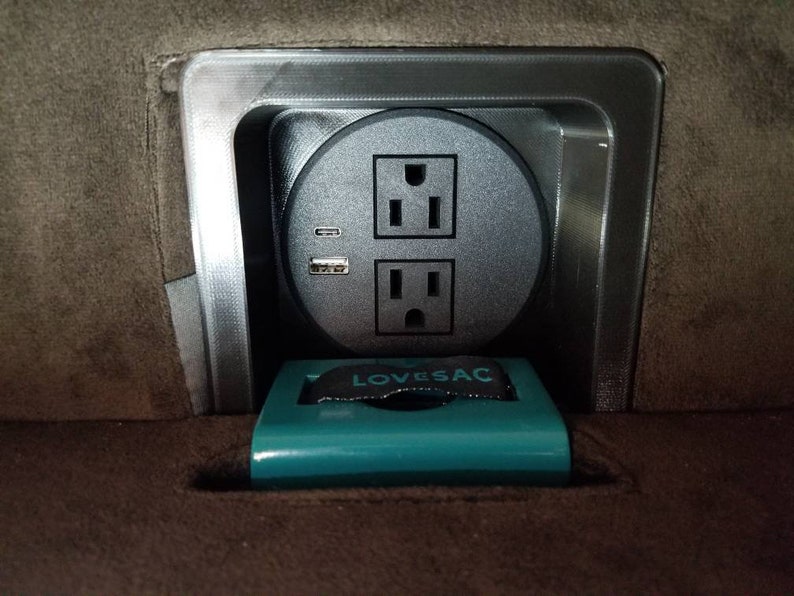 May include: A close-up of a grey electrical outlet with two standard sockets and a USB port. The outlet is mounted in a grey metal frame and is surrounded by a brown fabric surface. The text "LOVE SAC" is visible on a teal-coloured object in the foreground.