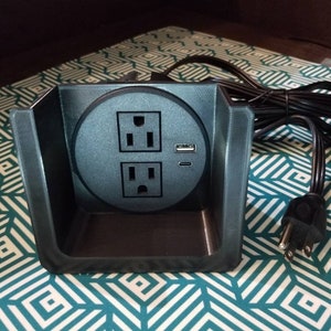 May include: A black electrical outlet with two standard sockets and a USB port. The outlet is mounted in a black plastic housing and has a black cord.
