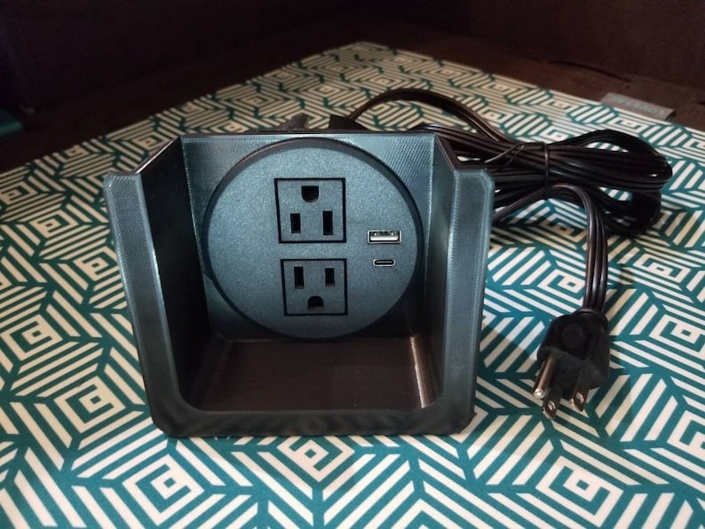 May include: A black electrical outlet with two standard sockets and a USB port. The outlet is mounted in a black plastic housing and has a black cord.