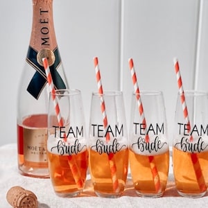 May include: Four clear glass champagne flutes with orange and white striped straws. Each flute is filled with a light orange beverage and has a black decal that reads "Team Bride".