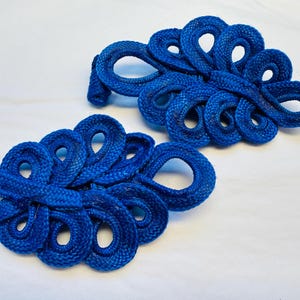 Op de afbeelding: Twee blauwe knopen van stof met een Chinees knoopontwerp, ook wel bekend als kikkergespen, met een decoratief, ingewikkeld ontwerp. Ze worden vaak gebruikt als versiering op kleding of accessoires.