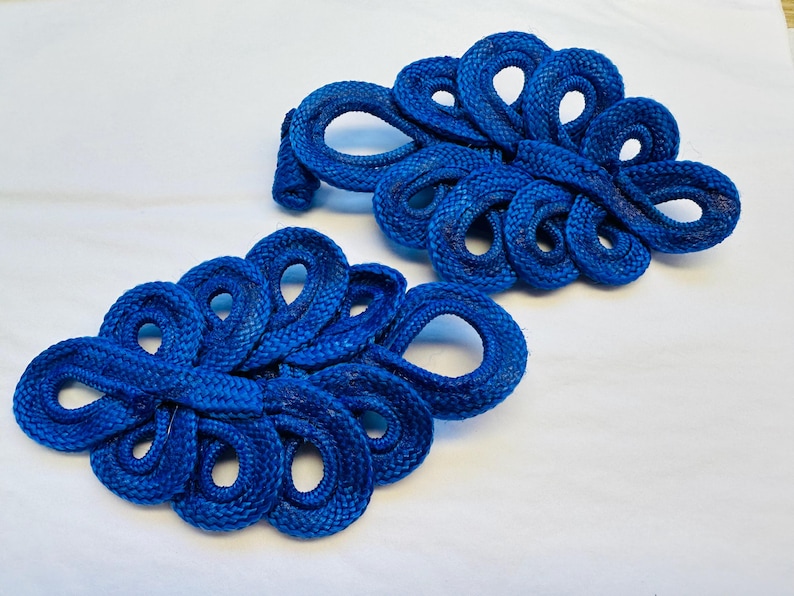 Op de afbeelding: Twee blauwe knopen van stof met een Chinees knoopontwerp, ook wel bekend als kikkergespen, met een decoratief, ingewikkeld ontwerp. Ze worden vaak gebruikt als versiering op kleding of accessoires.