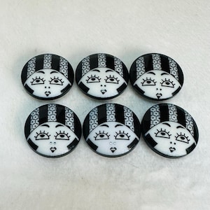 以下が含まれることがあります： 大きな目と小さな口をした女性の顔のスタイリッシュな画像が描かれた、黒と白の丸いボタンが6つ。ボタンには黒と白の幾何学模様が施されています。