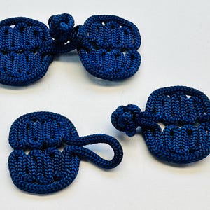 May include: A collection of four navy blue frog closures, a type of button, made from tightly woven cord. The closures feature a looped design and a knotted button. These are often used on jackets and coats.