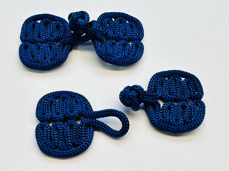 May include: A collection of four navy blue frog closures, a type of button, made from tightly woven cord. The closures feature a looped design and a knotted button. These are often used on jackets and coats.