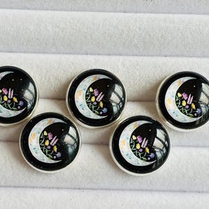 May include: Five circular buttons with a silver-toned metal rim. Each button features a black background with a crescent moon graphic filled with small, colourful floral illustrations in shades of yellow, purple, and green, set under a glossy finish.