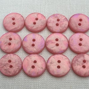 May include: A collection of twelve pink, round buttons, each with two holes, arranged in a grid. The buttons have a pearlescent finish with a marbled effect, showing shades of purple and white. They are set on a textured, off-white fabric.
