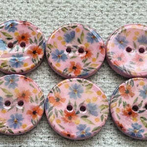 Puede incluir: Un conjunto de seis botones redondos de dos agujeros con una base de color rosa claro y un patrón estampado de pequeñas flores azules y naranjas, hojas verdes y diminutos puntos amarillos. Cada botón tiene un acabado liso y brillante.