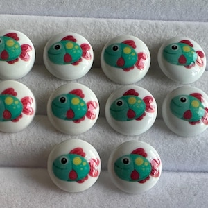 Può includere: Dieci pomelli per mobili in ceramica bianca con un design a pesce colorato. Ogni pomello presenta un pesce turchese con macchie gialle e pinne rosse, su sfondo bianco. I pomelli sono rotondi e adatti ai mobili.