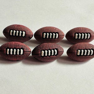 Puede incluir: Seis botones decorativos marrones en forma de balón de fútbol con detalles de costura blancos están dispuestos sobre una superficie blanca. Los botones tienen una superficie texturizada y están espaciados uniformemente.