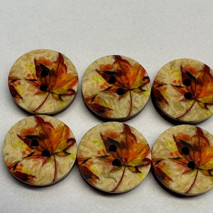 May include: Six circular wooden buttons with a fall leaf design. Each button features a maple leaf in shades of orange, yellow, and red against a cream background. The buttons have two black holes for sewing.